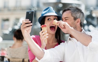 Portrait of a smiling couple eating ice cream and having fun in the city. The grey hair man with a beard in a white shirt is taking a picture with phone. The woman is wearing a blue hat and a pink top. They are making faces with their ice cream.